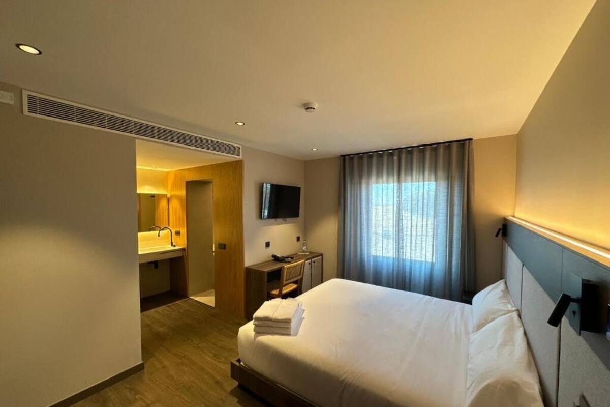 A hotel room with a large bed, a desk, a chair, a TV, and a bathroom with a sink and mirror.
