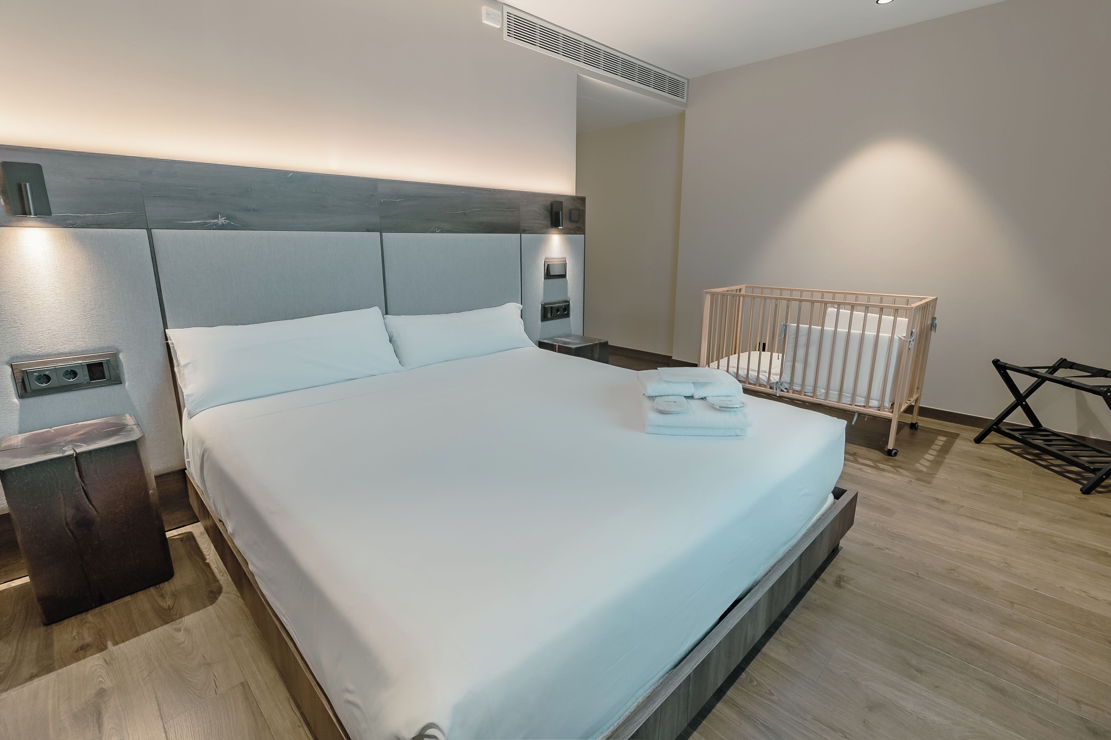Habitación de hotel moderna con una cama grande, cabecera de madera, una mesita de noche, una cuna y una pequeña mesa con toallas dobladas.