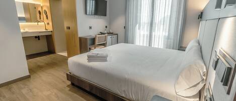 Una habitación de hotel moderna con una cama grande, un escritorio con silla, un televisor montado en la pared y un baño con lavamanos y espejo.
