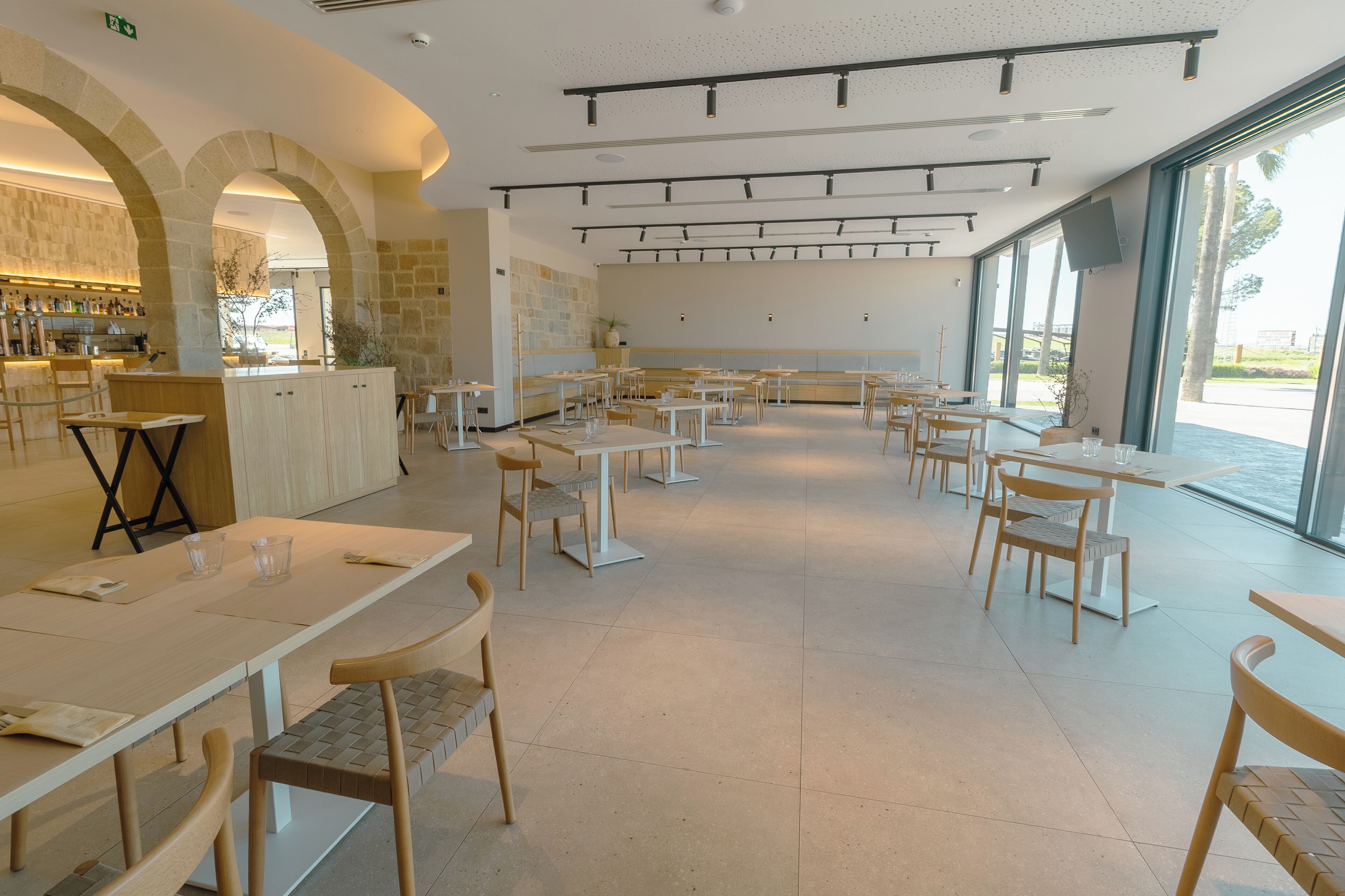 A spacious dining area with multiple tables and chairs, a bar area with shelves of bottles, and large windows offering a view of the outdoors.