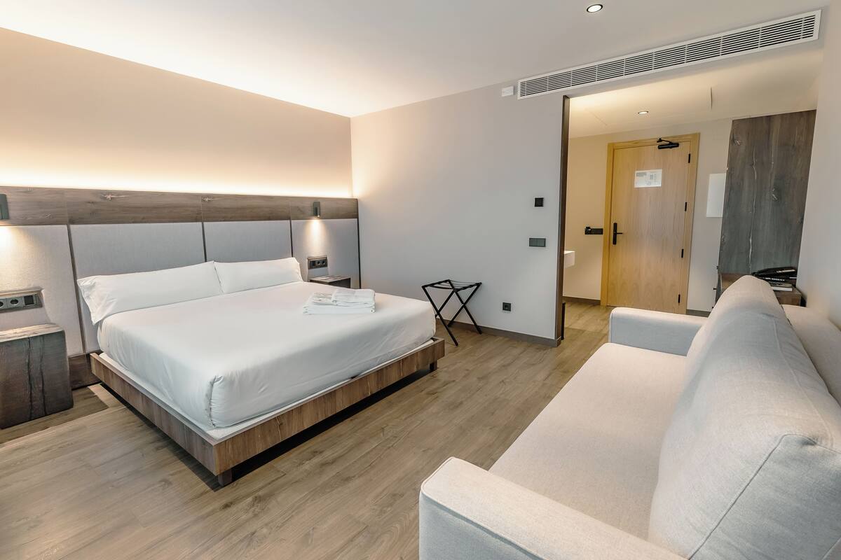 A modern hotel room with a large bed, a sofa, and a wooden floor.
