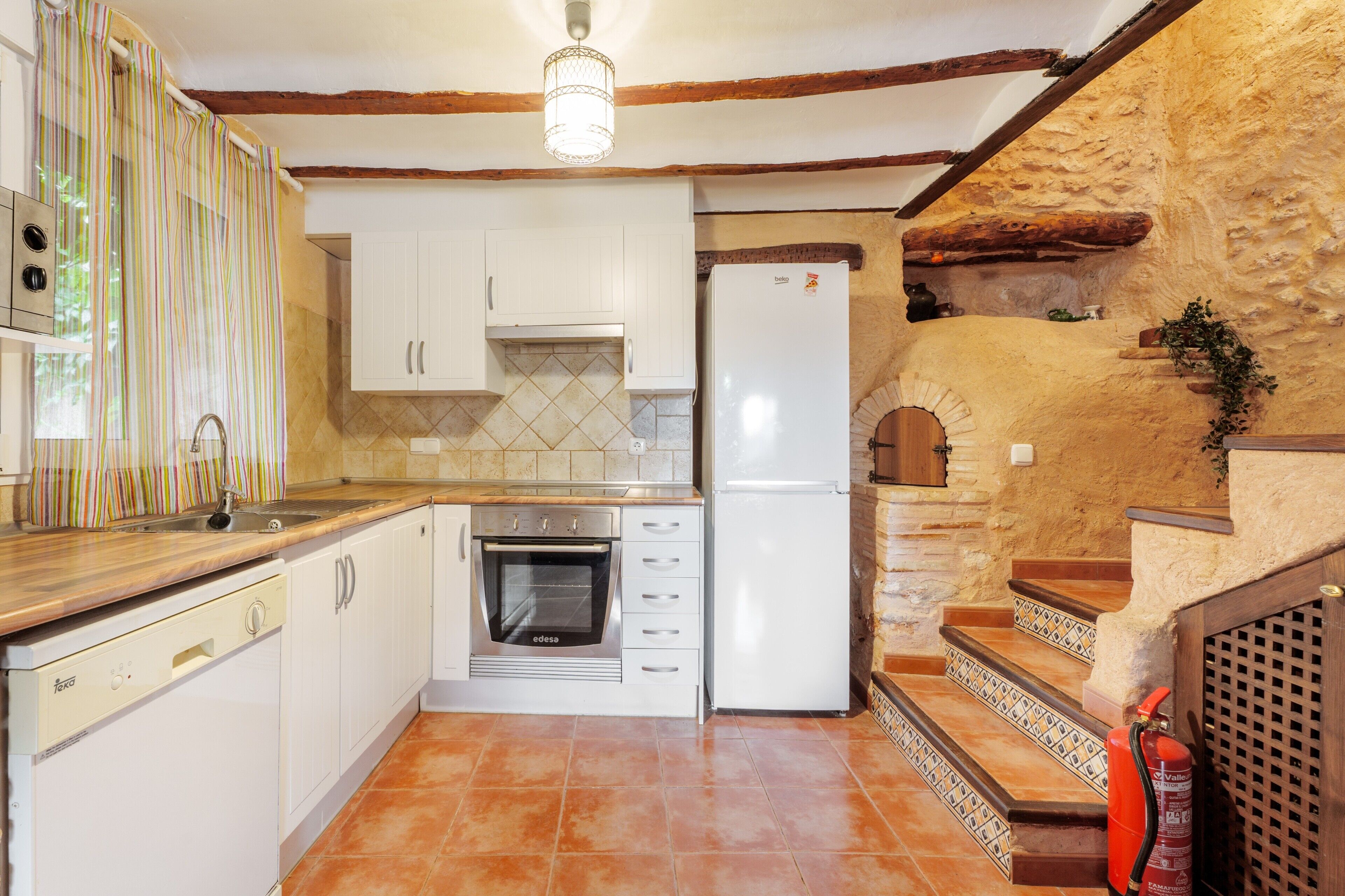 Cuina amb armaris blancs, forn de bola, nevera i escala de fusta que condueix a un nivell superior.