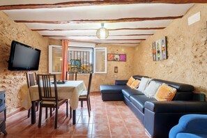 A room with a dining table, chairs, a sofa, a television mounted on the wall, and a window with curtains.