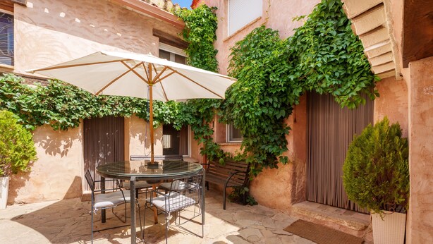 A stone-floored patio with a glass table and chairs, an umbrella, a bench, and a door with a wooden shutter.