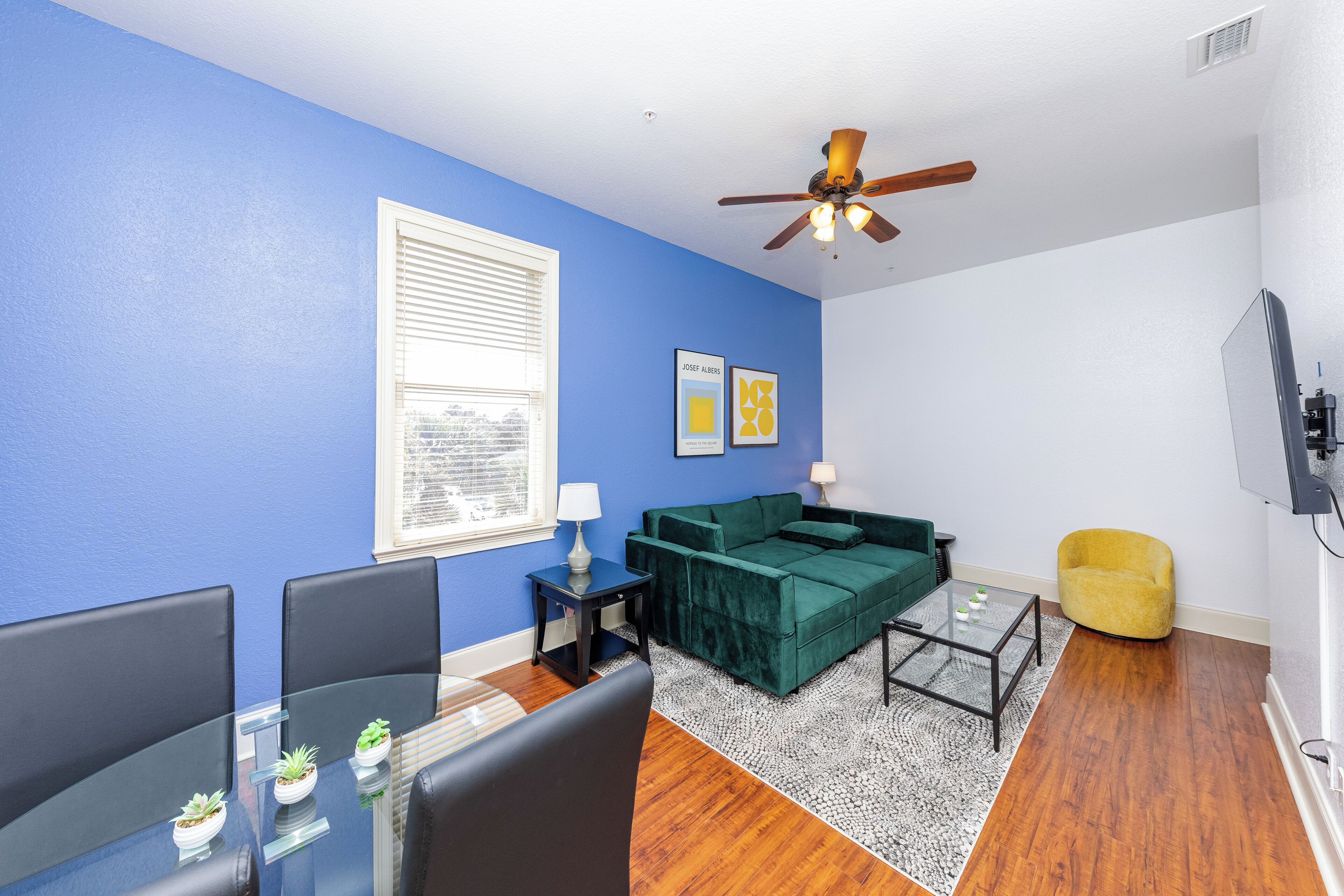 A living room with a blue wall, a green sofa, a yellow armchair, a glass coffee table, and a ceiling fan.