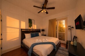 A hotel room with a bed, a ceiling fan, a bedside lamp, a window with curtains, a wall-mounted TV, and a bedside table with a remote control.