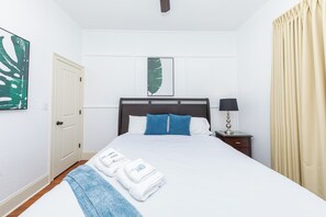 Una cama bien hecha con sábanas blancas y almohadas azules, una mesita de noche con una lámpara y una puerta a la izquierda.