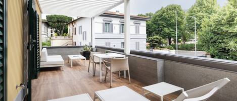 A covered outdoor seating area with white furniture, a view of a building with shutters, and greenery in the background.