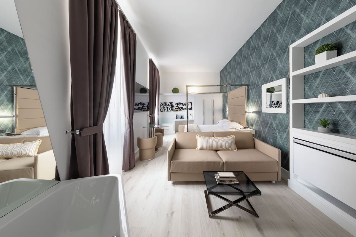 A modern hotel room with a beige sofa, a glass coffee table, a bed with a large mirror, and a wall with a geometric pattern.