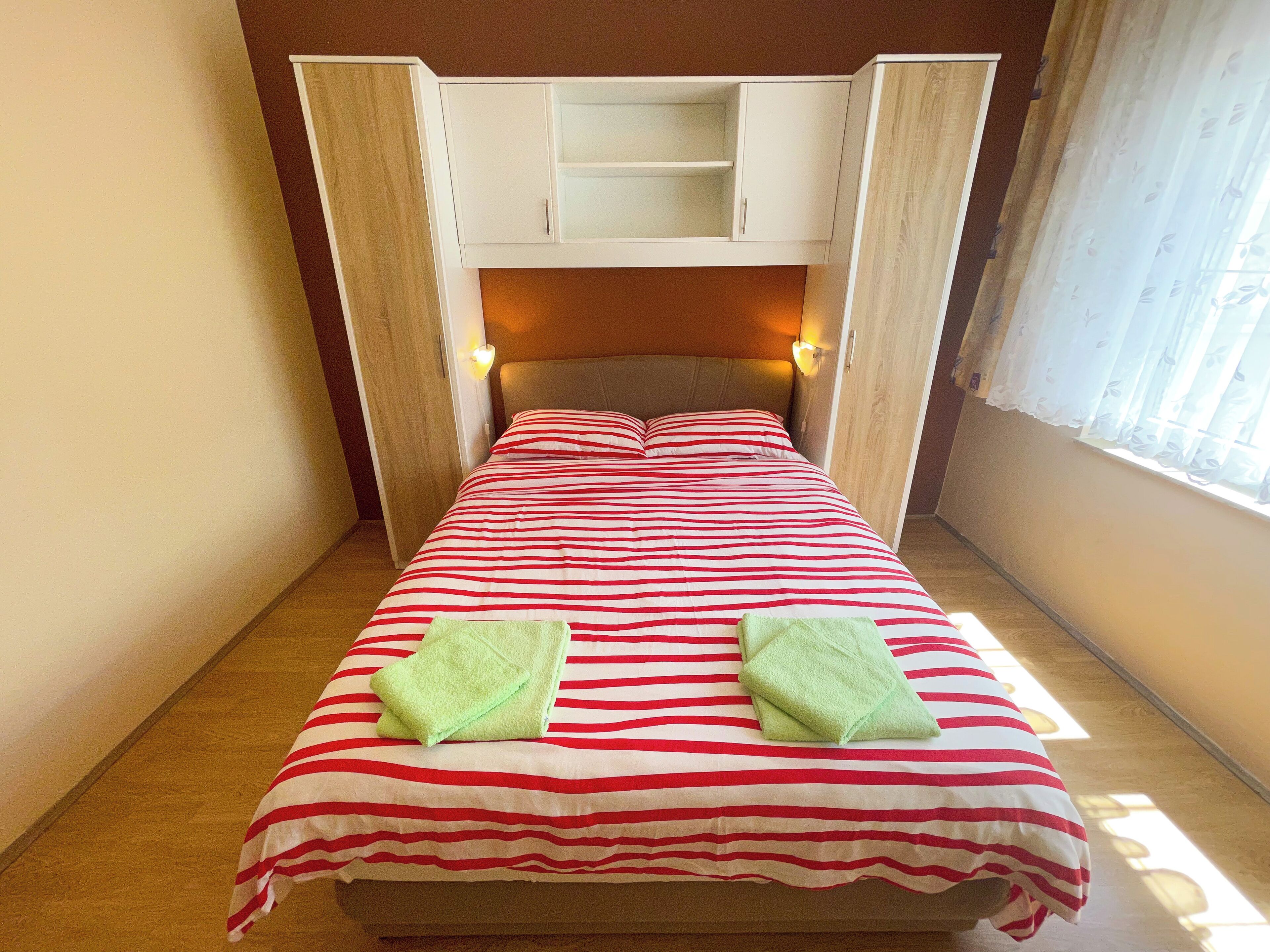 A bed with a striped bedspread, two green towels, a headboard with built-in lighting, and a window with sheer curtains.