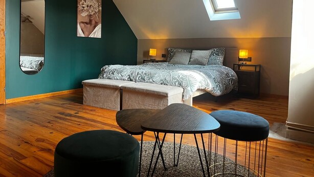 A bedroom with a sloped ceiling, a skylight, a bed with patterned bedding, a bench, two stools, a round table, and a large mirror.