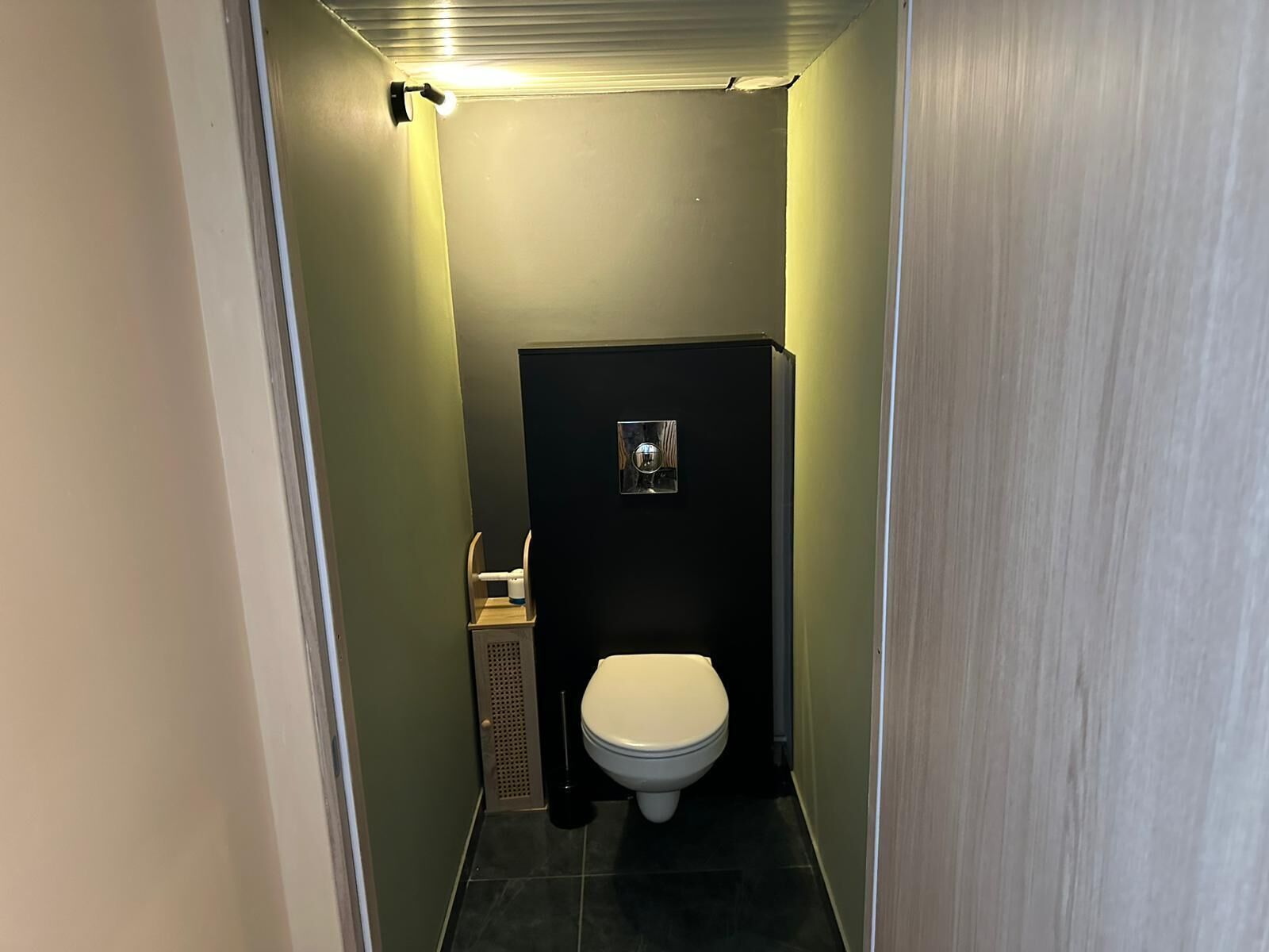 A narrow hallway leading to a small, dark-colored bathroom with a toilet, a trash bin, and a wall-mounted light fixture.
