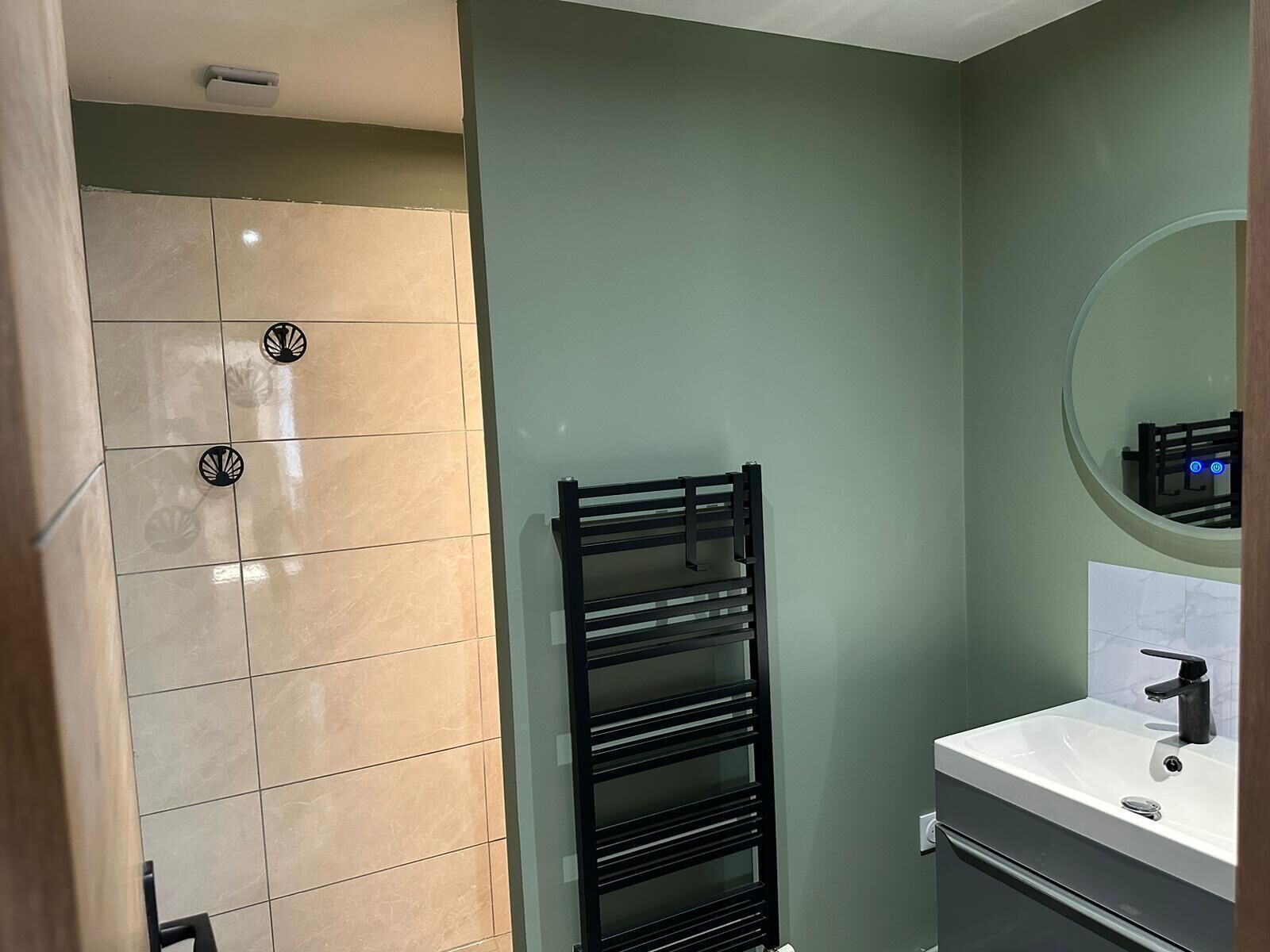 Une salle de bain avec des carreaux de couleur beige, un porte-serviettes noir, un lavabo blanc avec un robinet noir et un miroir au-dessus du lavabo.