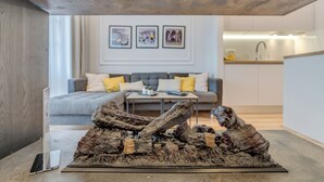 A modern living room with a grey sofa, a coffee table, and a fireplace with a log centerpiece. There is a kitchen in the background with white cabinets and a sink. - Romanca Deluxe Studio - City View (Split)