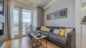 A modern living room with a grey sofa, a coffee table, and a large window with curtains. There are two framed pictures on the wall and an air conditioning unit above the sofa. - Romanca Deluxe Studio - City View (Split)