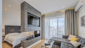A modern hotel room with a large bed, a sofa, a flat-screen TV mounted on a stone wall, and a large window with curtains. - Romanca Deluxe Studio - City View (Split)