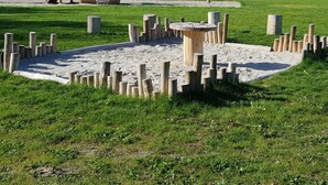 Un parque infantil con un tobogán de madera y estructuras para escalar, rodeado de arena y zonas verdes.