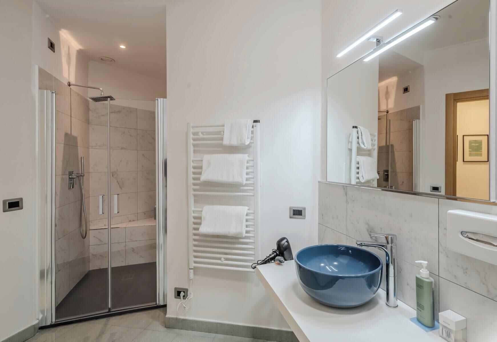 Un bagno moderno con cabina doccia in vetro, uno specchio grande, un lavandino blu e un portasciugamani.