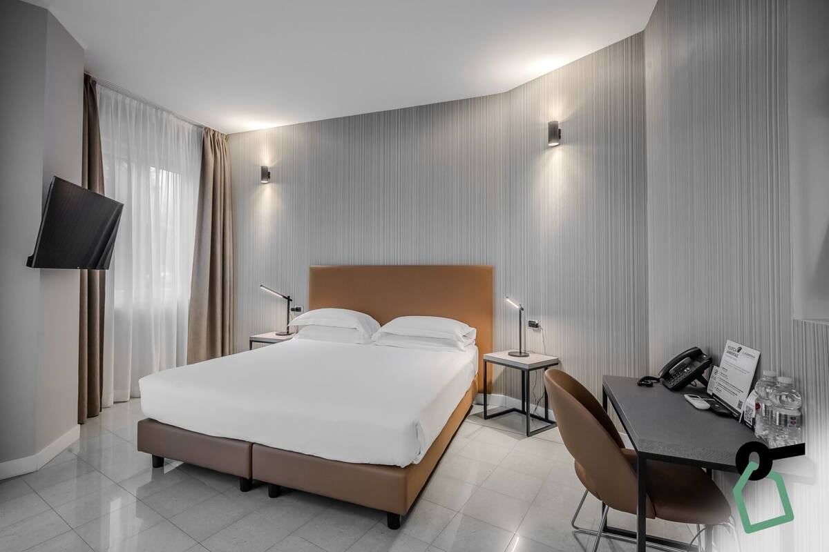 A modern hotel room with a large bed, bedside tables, a desk with a chair, a flat-screen TV, and a telephone.