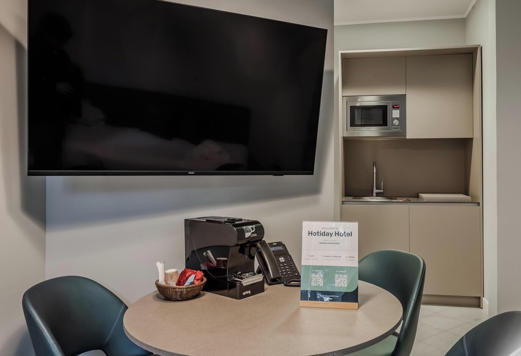 A hotel room with a flat-screen TV, a coffee machine, a sign for the Holiday Hotel, and a kitchenette with a microwave.