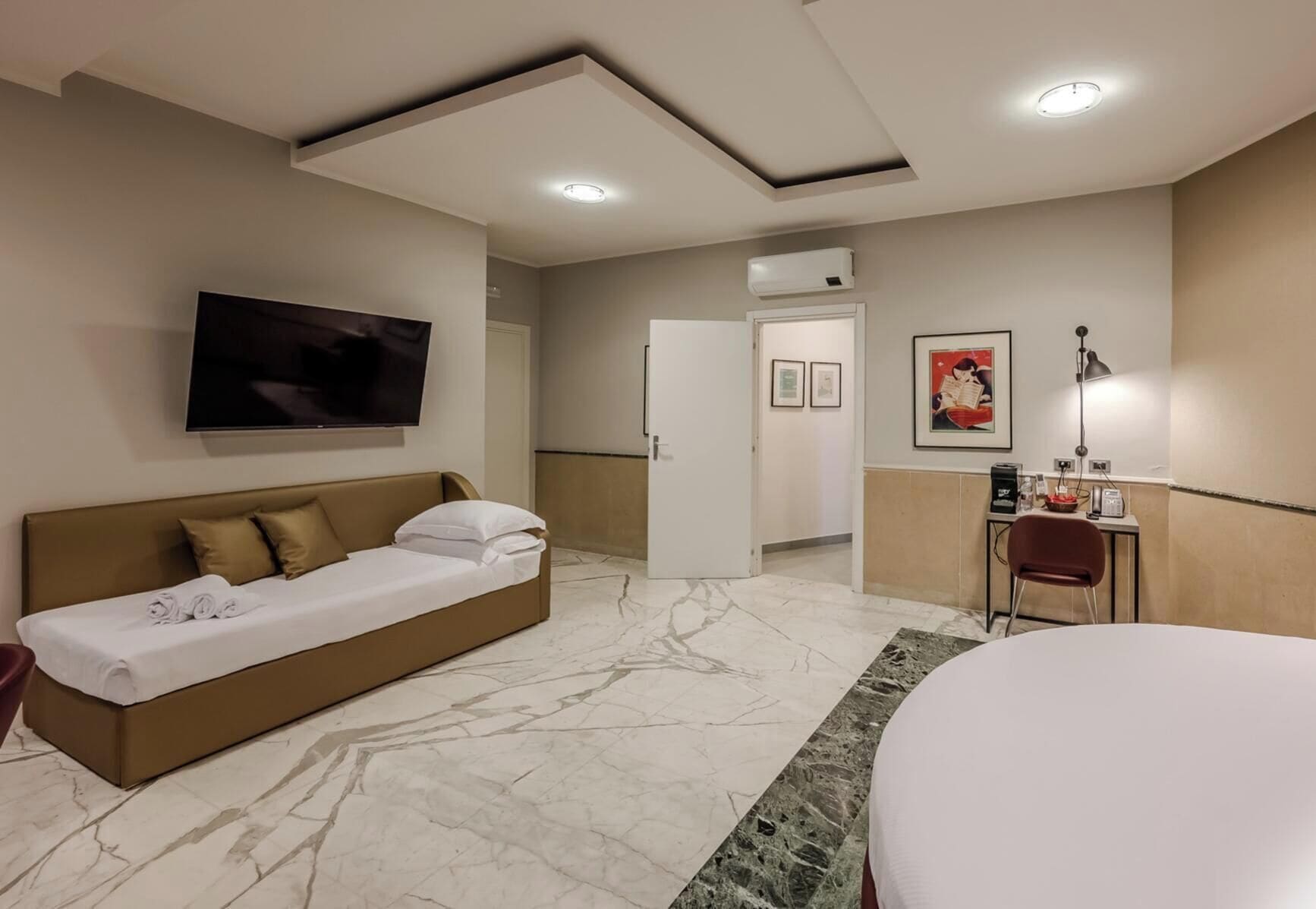 Ein modernes Hotelzimmer mit einem Bett, einer Schlafcouch, einem Schreibtisch mit Stuhl, einem Flachbildfernseher und Kunstwerken an den Wänden.