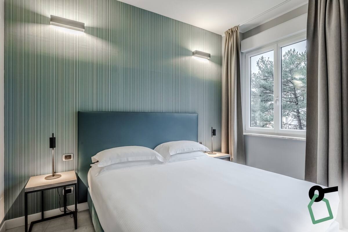 A hotel room with a bed, a nightstand, a lamp, and a window with curtains.