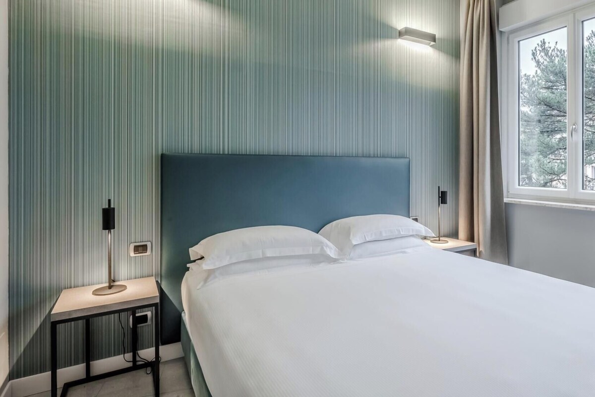 Ett hotellrum med en säng, ett nattduksbord, en lampa och ett fönster med gardiner.
