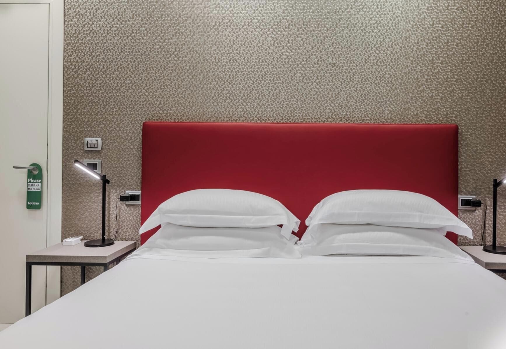 A hotel room with a red headboard, two bedside tables with lamps, and a bed with white bedding.