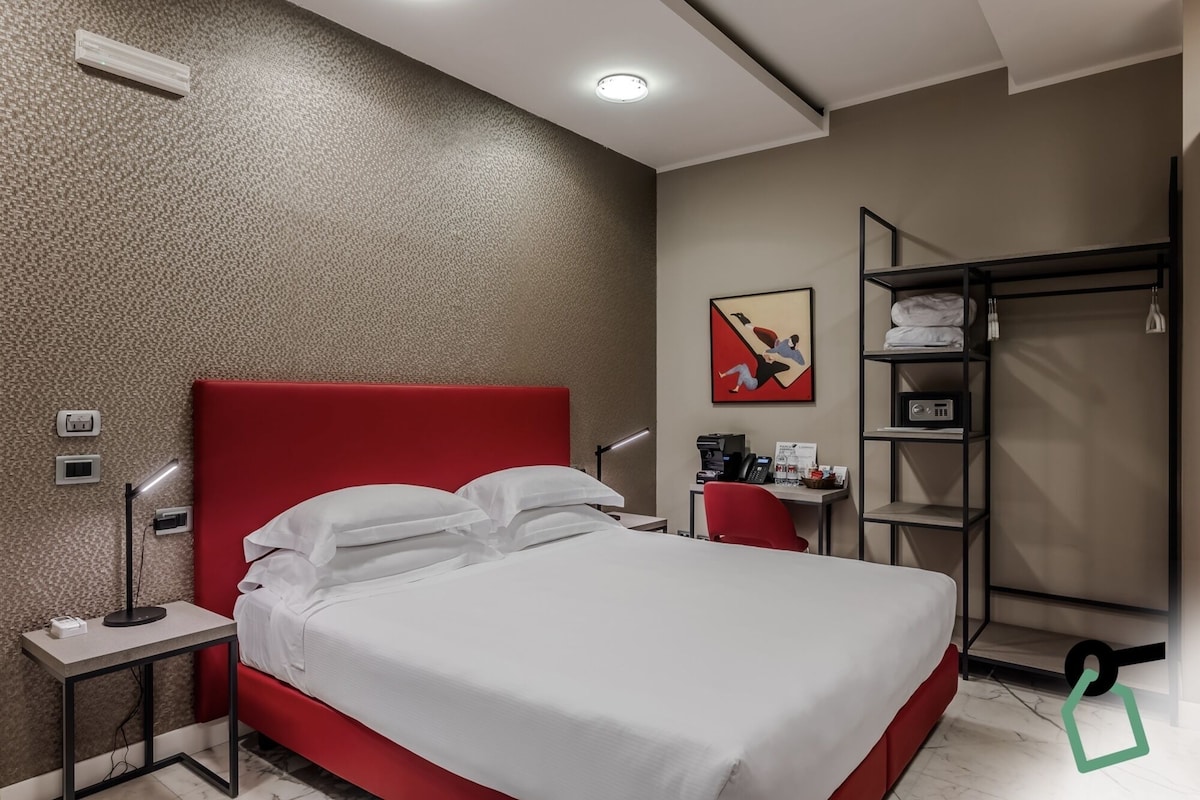 A hotel room with a red headboard, a bed with white linens, a desk with a computer, a red chair, a coat rack, and a painting on the wall.