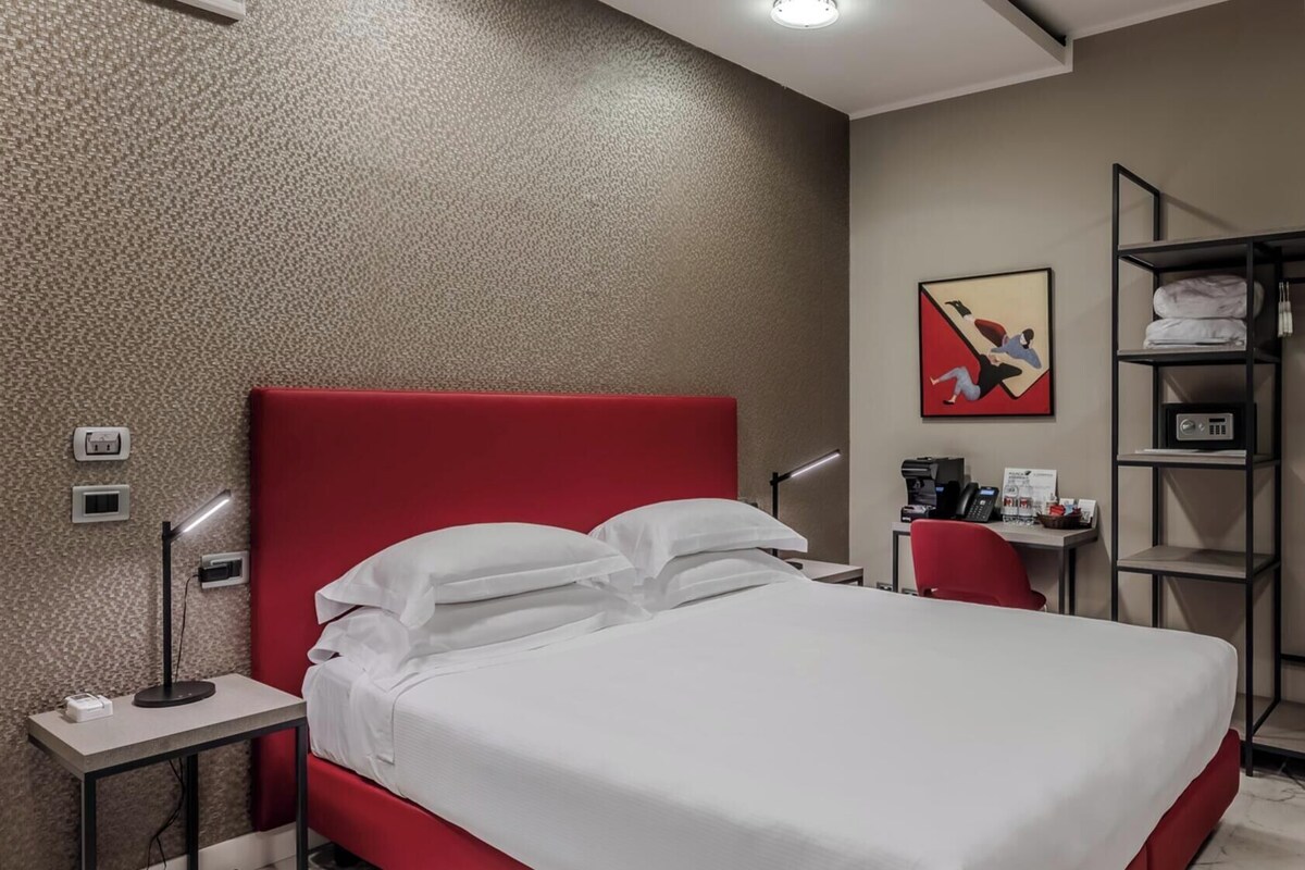 Ett hotellrum med en röd sänggavel, en säng med vita sängkläder, ett skrivbord med en dator, en röd stol, en klädställning och en tavla på väggen.