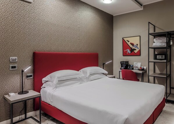 A hotel room with a red bed, a desk with a lamp, a shelf with towels, and a painting on the wall. - Hotiday Apartments La Spezia (La Spezia)