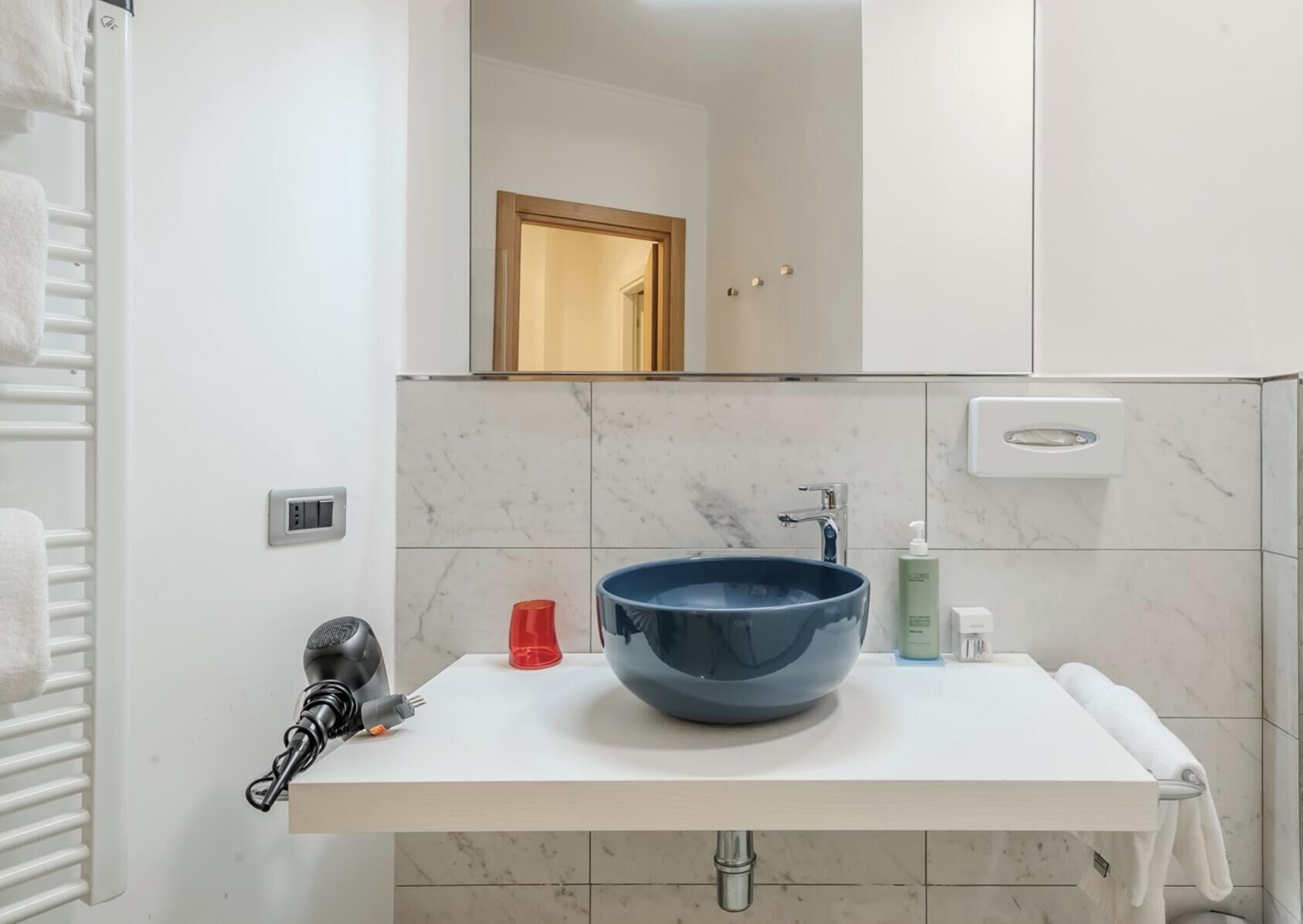 Une salle de bain avec un lavabo blanc, un récipient bleu, un gobelet rouge, une bouteille verte et un porte-serviettes.