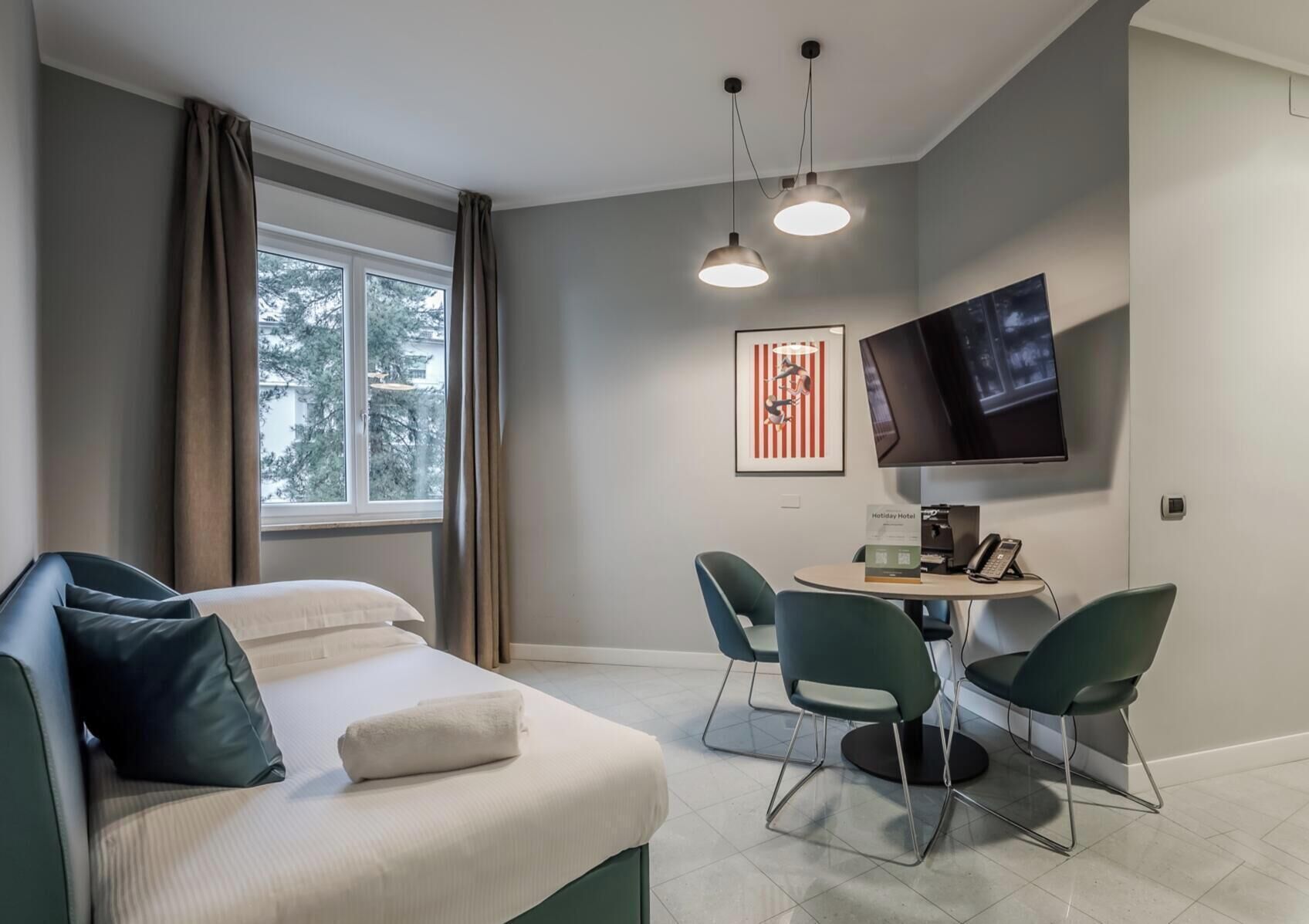 A hotel room with a bed, a desk with chairs, a flat-screen TV, and a window with curtains.