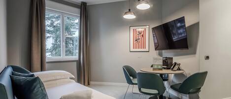 A hotel room with a bed, a desk with chairs, a flat-screen TV, and a window with curtains.