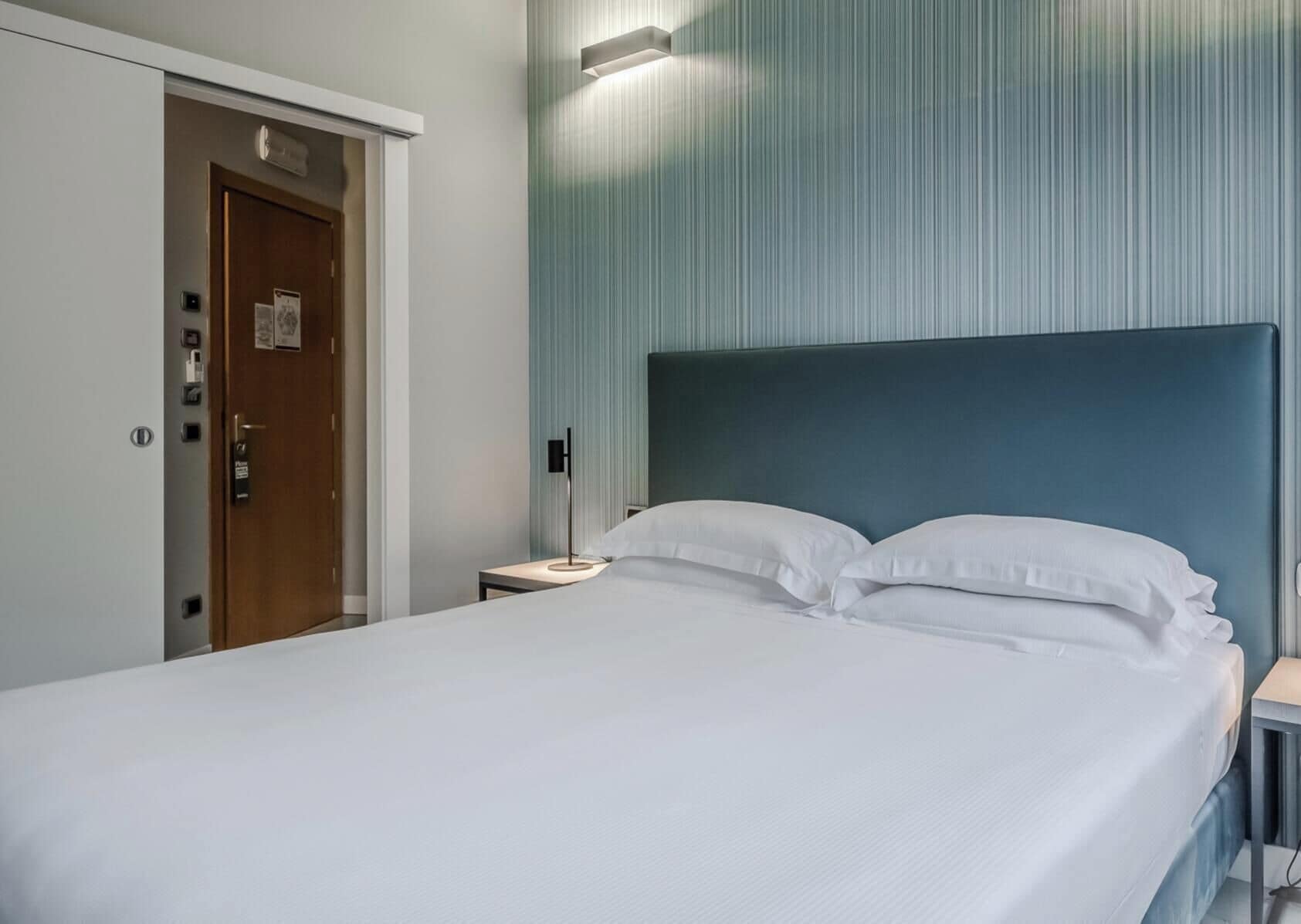 Una camera d'albergo con un letto, un comodino, una porta e un punto luce fissato al muro.