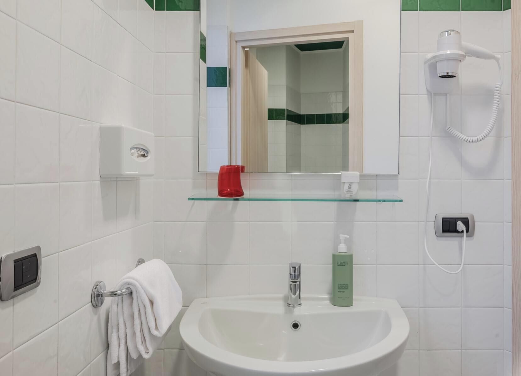 Une salle de bain avec un lavabo blanc, un sèche-cheveux portatif, un gobelet rouge et une étagère en verre.