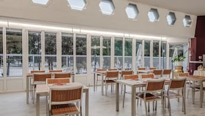 A spacious indoor dining area with multiple tables and chairs, large windows with a view of trees, and a ceiling with hexagonal skylights. - Hotiday Apartments La Spezia (La Spezia)
