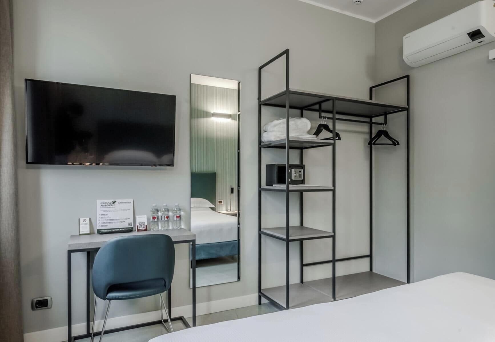 Una habitación de hotel con cama, escritorio con silla, televisión, armario con estantes y unidad de aire acondicionado.