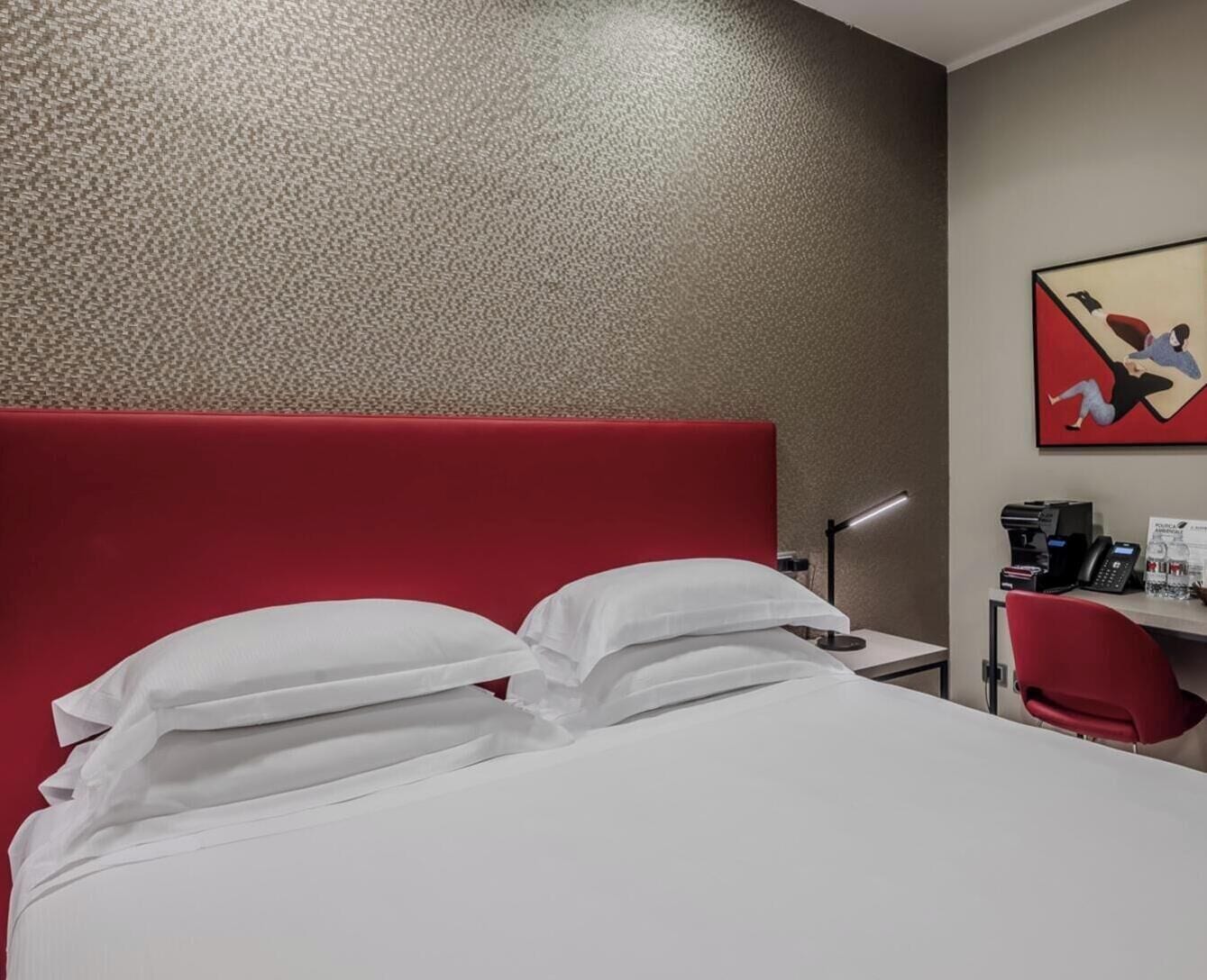 Habitación de hotel con cabecera roja, ropa de cama blanca, escritorio con lámpara y un cuadro en la pared.