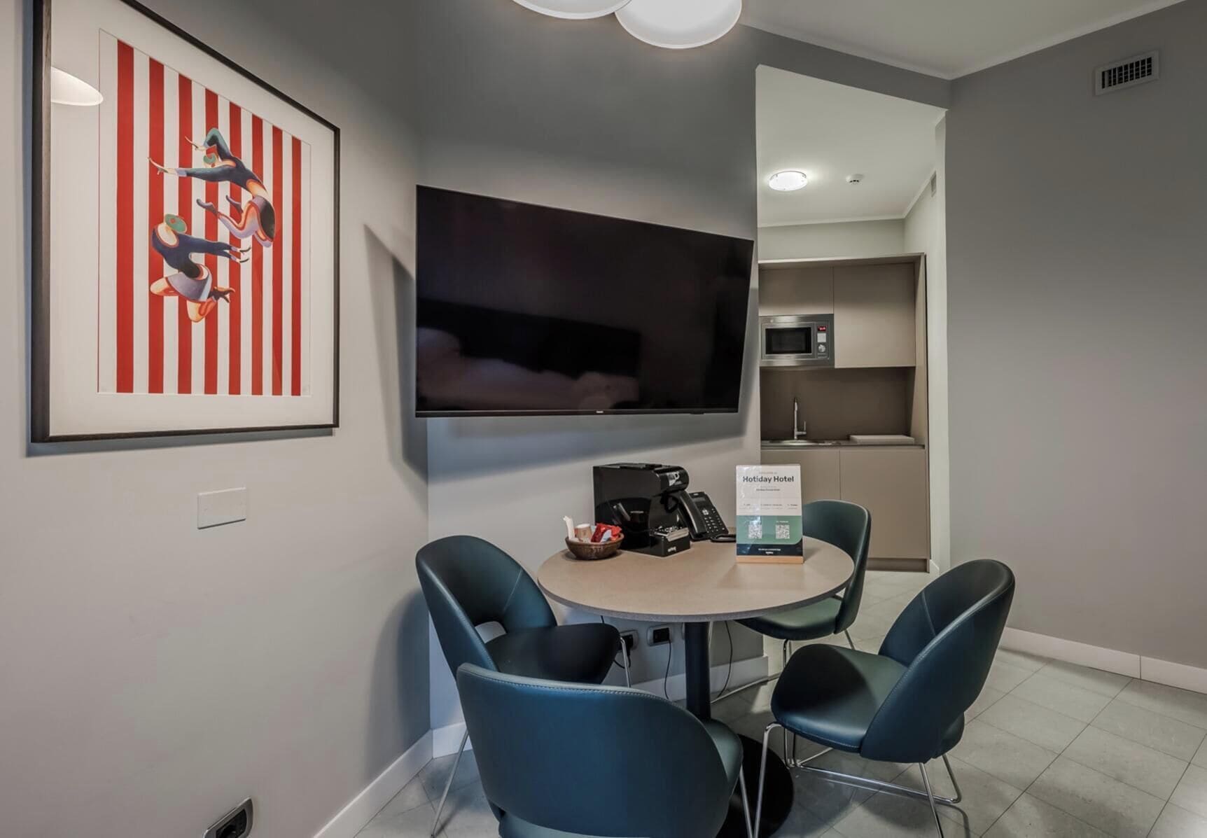 Habitación de hotel con televisor de pantalla plana, una pequeña mesa con sillas, una cafetera y una obra de arte enmarcada en la pared.