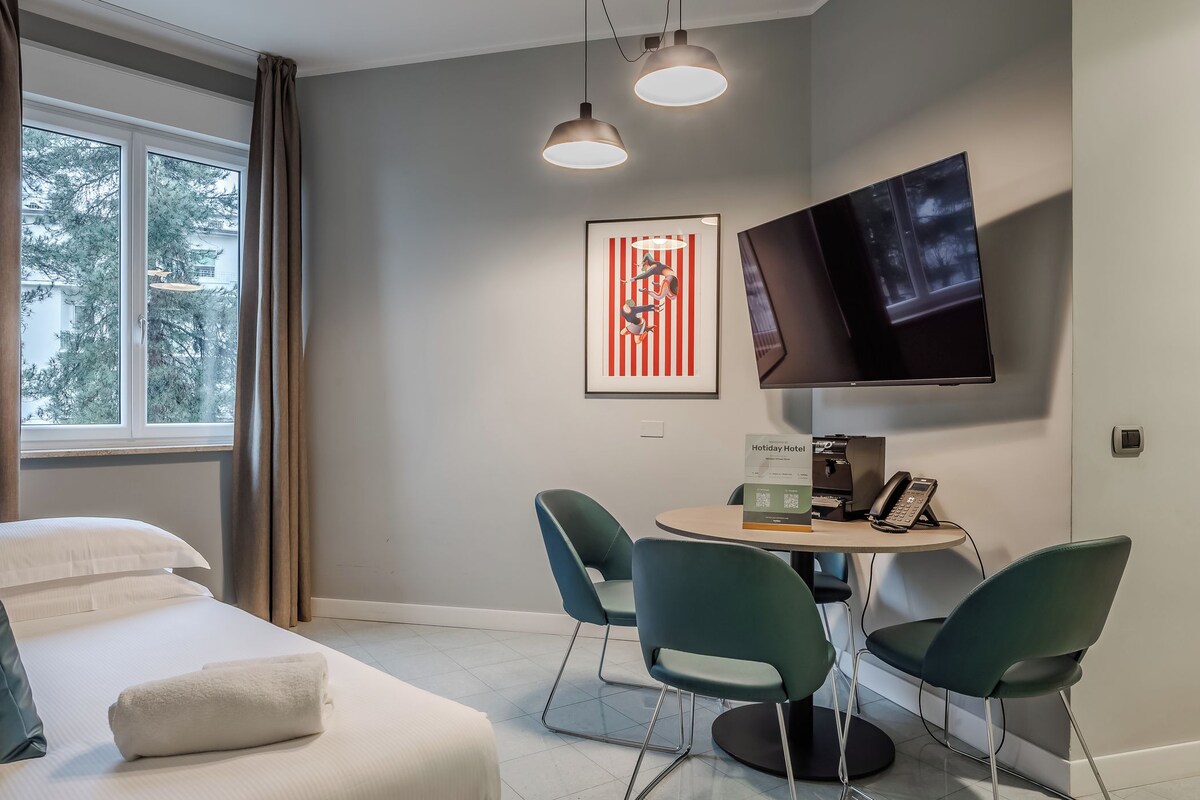 A hotel room with a bed, a desk with chairs, a flat-screen TV, a framed artwork, and a window with curtains.