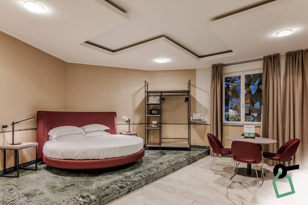 A hotel room with a round bed, a desk, a chair, a small table, a wardrobe, and a window with curtains.