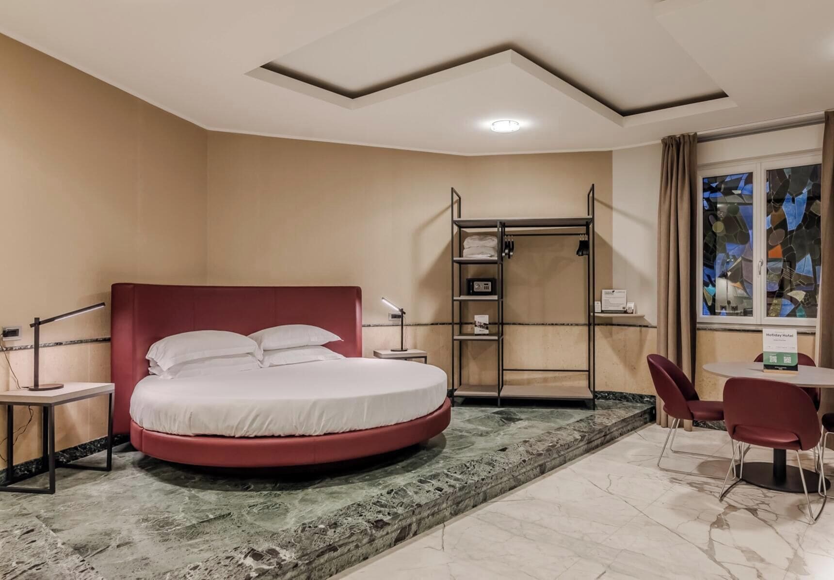 A hotel room with a circular bed, a walk-in closet, a desk with a chair, and a view of the outside through a window.