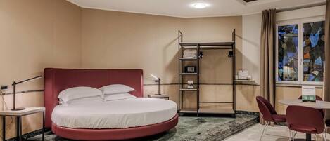 A hotel room with a circular bed, a walk-in closet, a desk with a chair, and a view of the outside through a window.