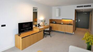 A modern hotel room with a wooden desk, a television, a chair, and a small kitchenette.