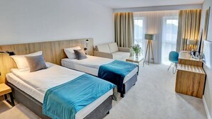 Egy szállodai szoba két ággyal, íróasztallal, székkel, televízióval és egy nagy, függönnyel ellátott ablakkal.