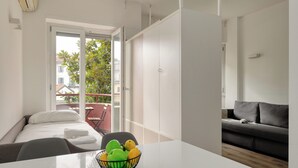 A modern hotel room with a bed, a dining table with a fruit bowl, a grey sofa, and a balcony with a view of buildings and trees.
