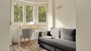 A modern room with a grey sofa, a small desk with a chair, and a window with a view of trees.