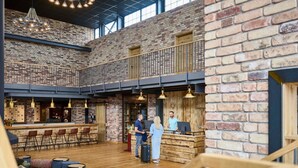 Ruime binnenruimte met een hoge plafond, openliggende bakstenen muren en een tussenverdieping. Er zijn zithoeken met tafels en stoelen, plus een bar met een counter en krukken.