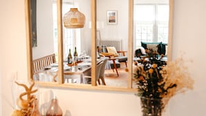 A dining area with a table set for two, a hanging light fixture, and a view of a living room through a mirror.
